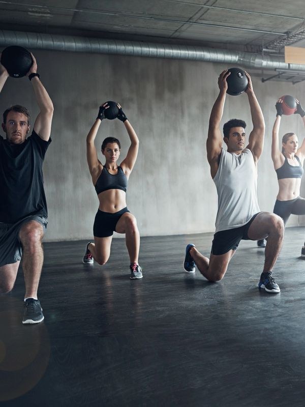 Four people exercising with medicine balls in a gym, performing lunges.