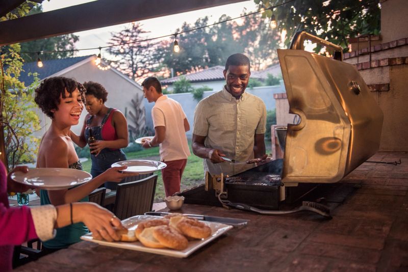 Multi ethnic hipsters enjoying backyard summer barbecue. The African American male who is manning the BBQ is serving the guests hamburgers.