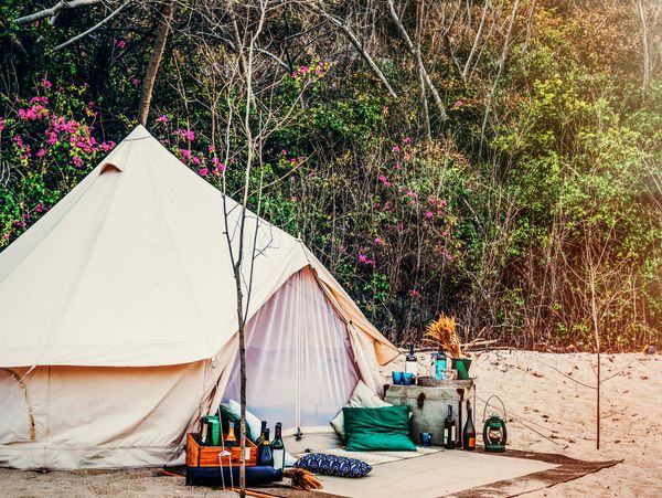 Cozy beige tent set up on sandy ground with colorful cushions and bottles, surrounded by trees with pink flowers.
