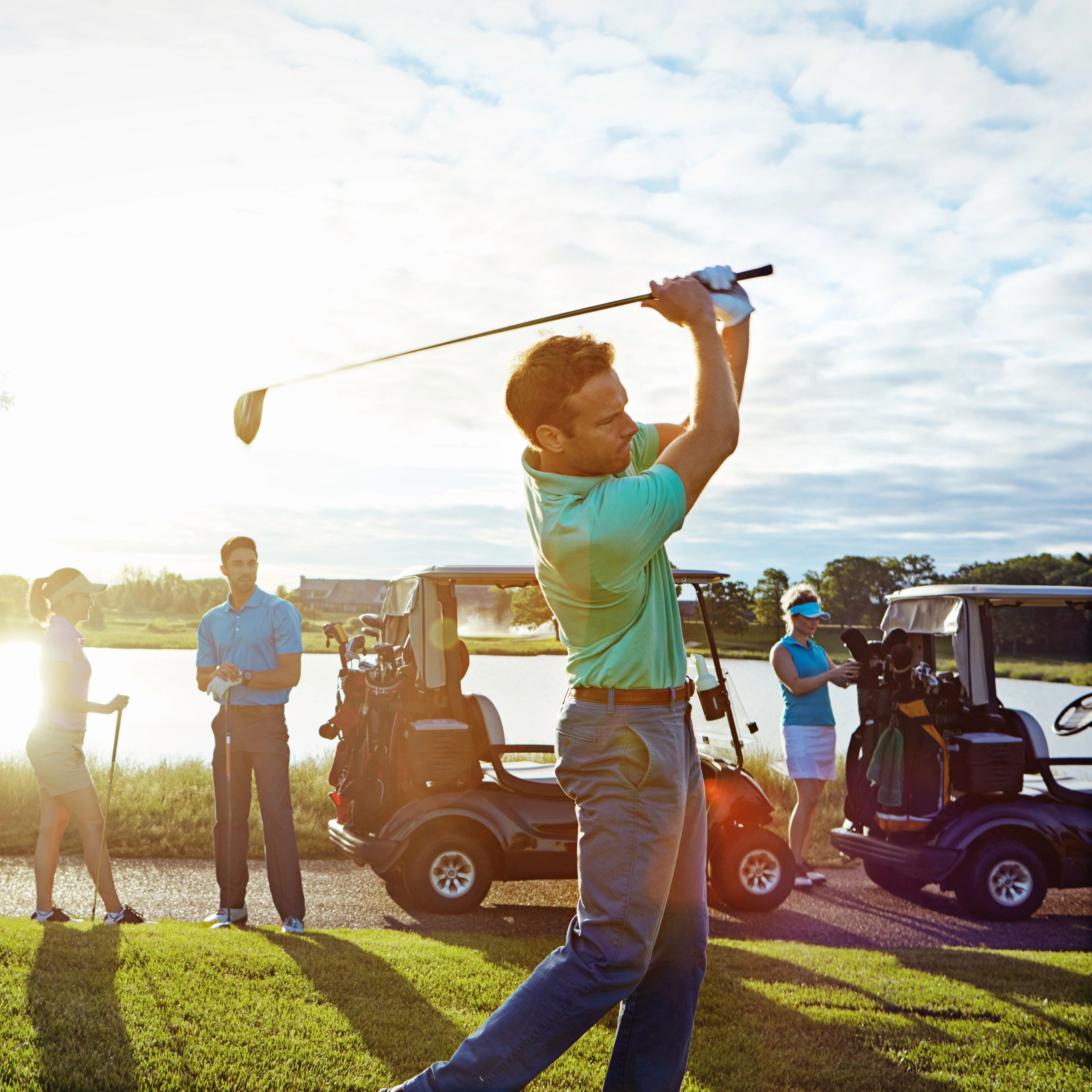 Man finishing a golf swing with friends and golf carts nearby.