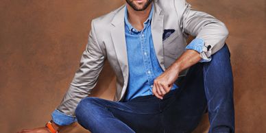 Stylish man in a light blazer, denim shirt, and brown shoes with blue laces sitting on the floor.