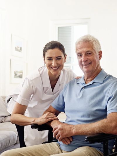 A nurse supports an elderly man in a wheelchair, both smiling warmly.