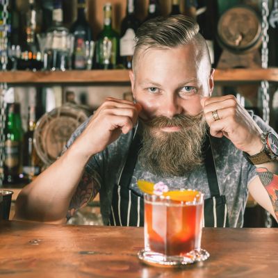 Bearded bartender adjusting his mustache behind a cocktail.