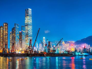 Night view of a vibrant city skyline with illuminated skyscrapers and cranes by the water.