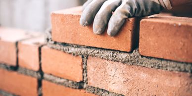 A gloved hand placing a brick on a wall under construction.
