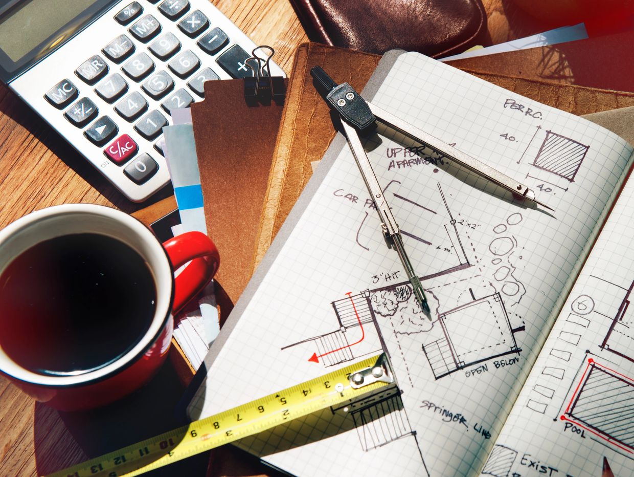 Architectural sketches with drawing tools, calculator, and coffee cup on a wooden desk.