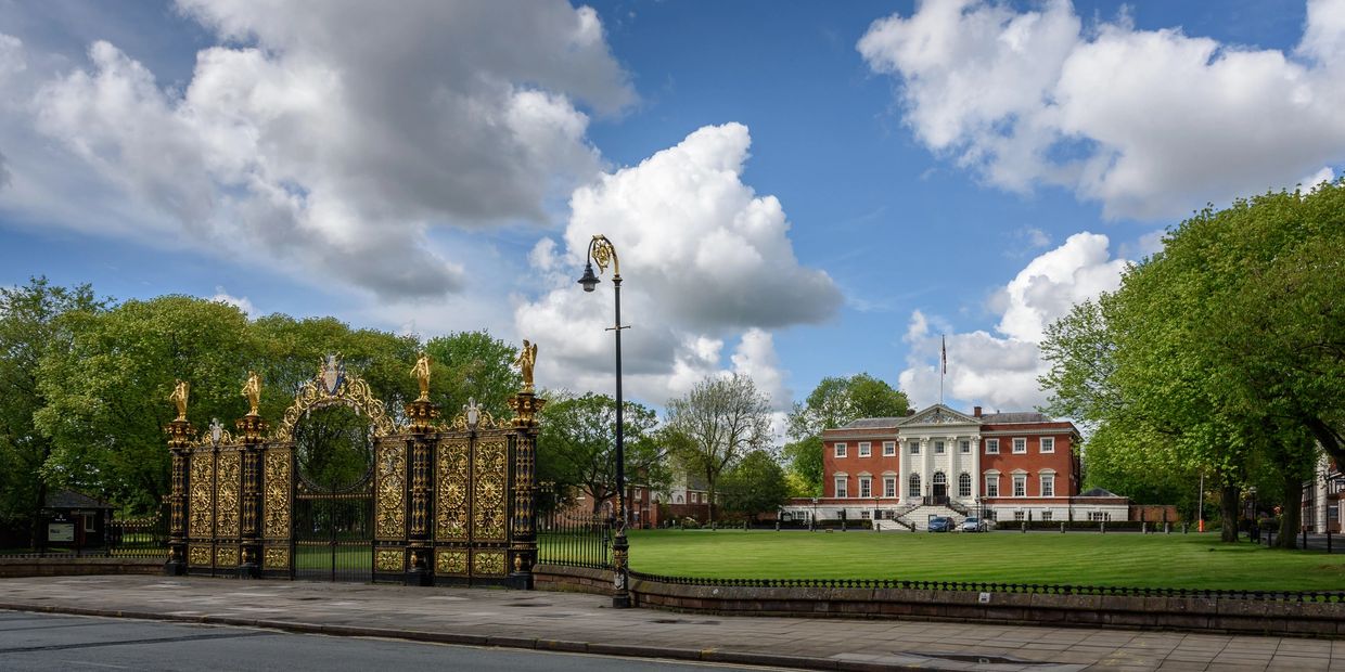 Ornate golden gates leading to a stately red-brick mansion with white columns under a partly cloudy sky.