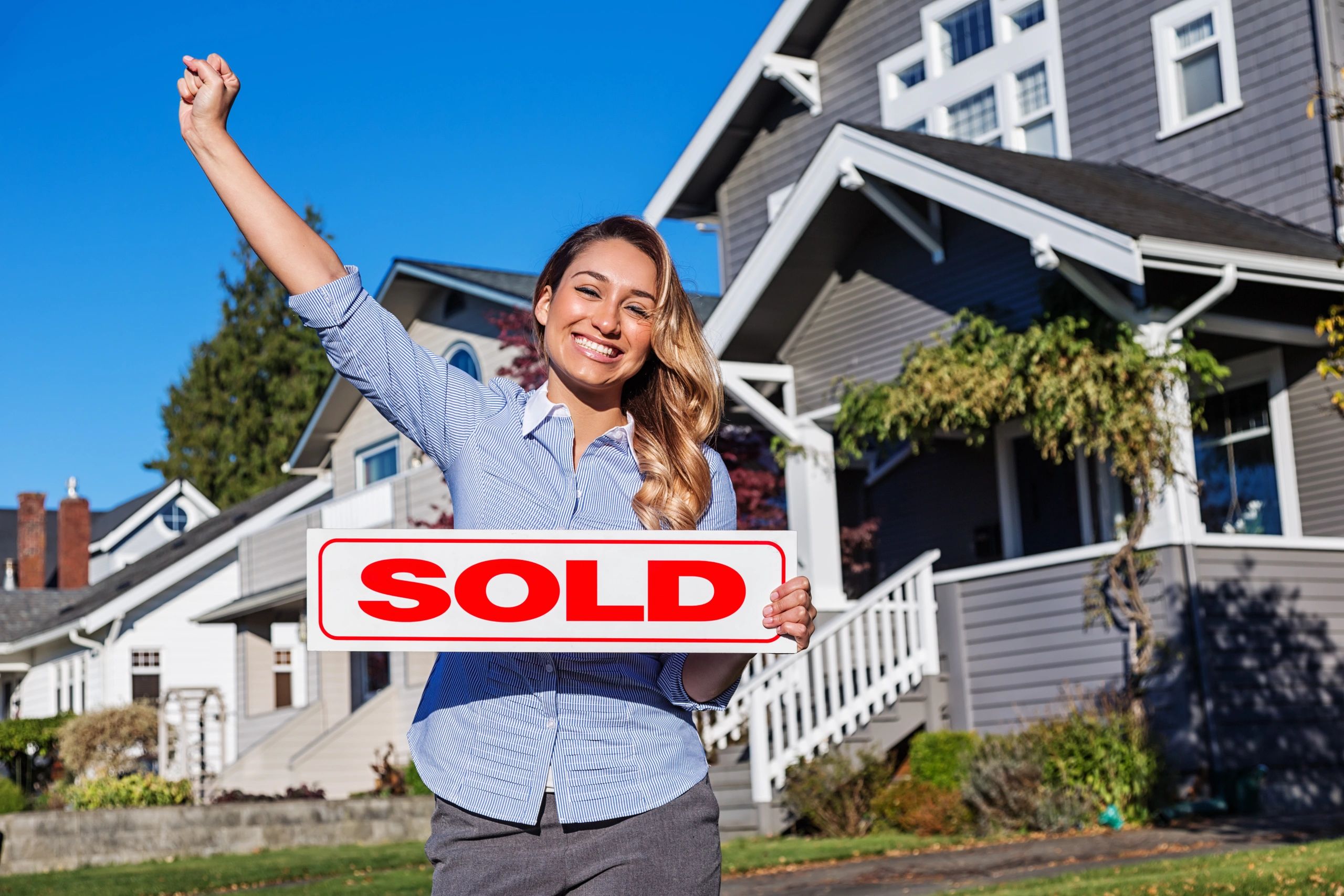 Happy woman holding a sold sign in front of a house on a sunny day.