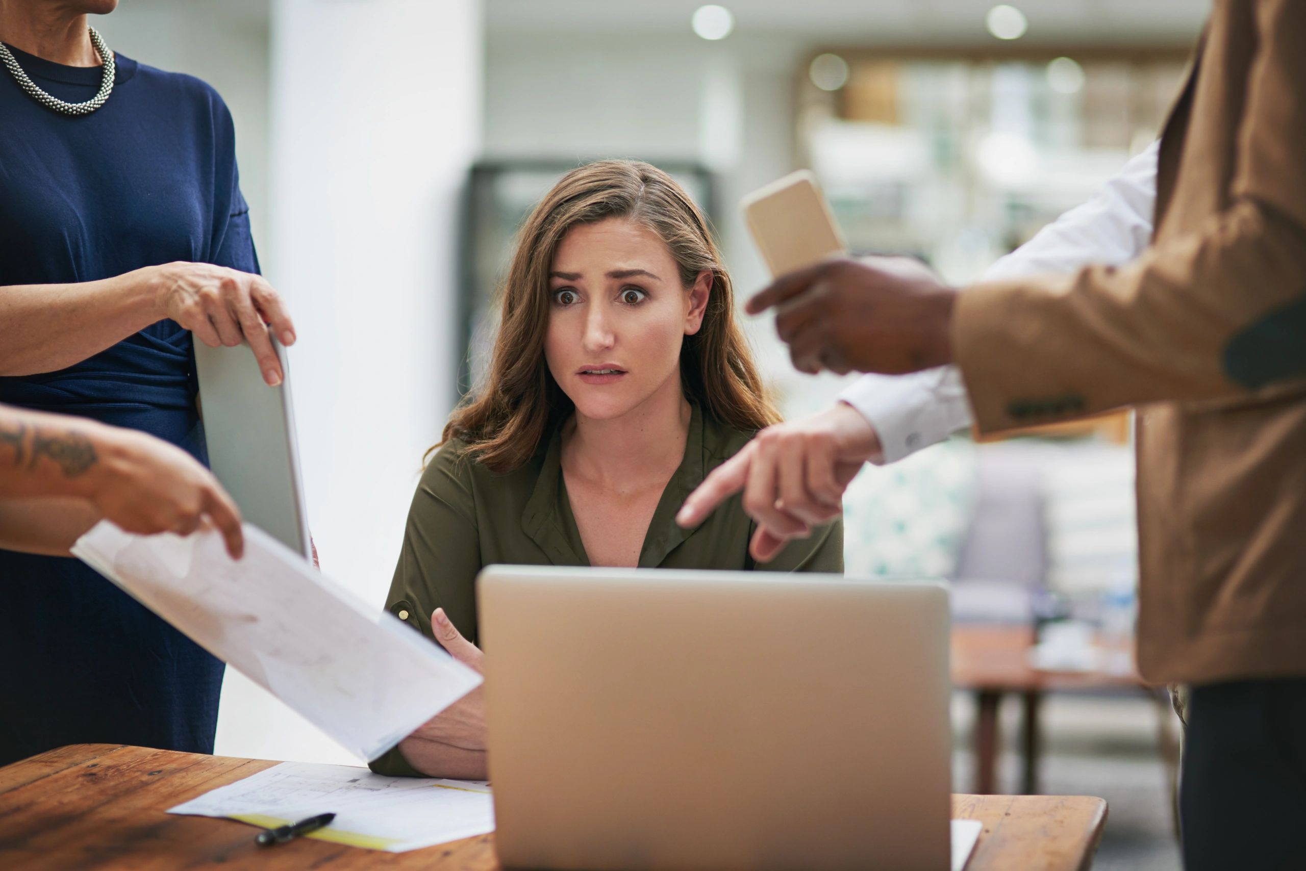 A woman looks stressed while colleagues point and show documents around her.