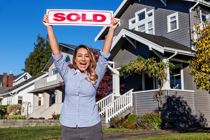 Photo of a cheerful young Hispanic businesswoman/Real Estate Agent with a red SOLD sign, standing in front of a row of suburban houses.