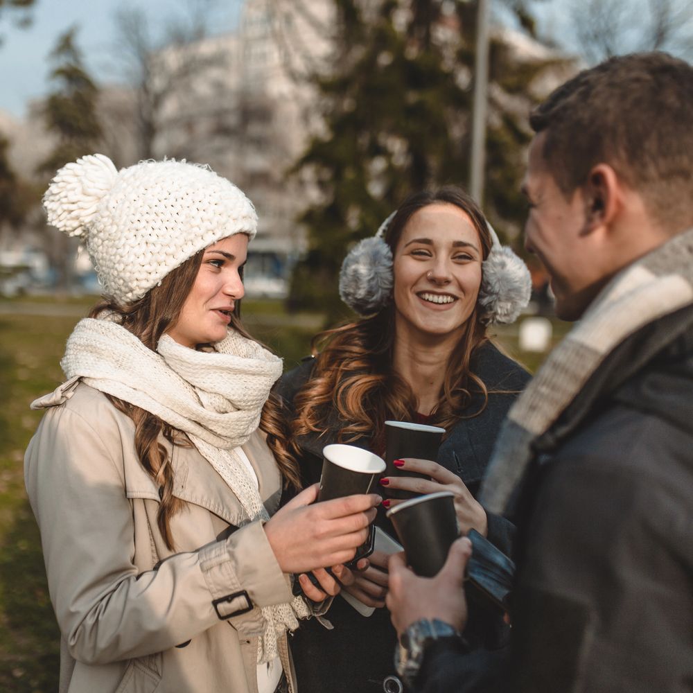 Three friends outdoors in winter, enjoying hot drinks and smiling.