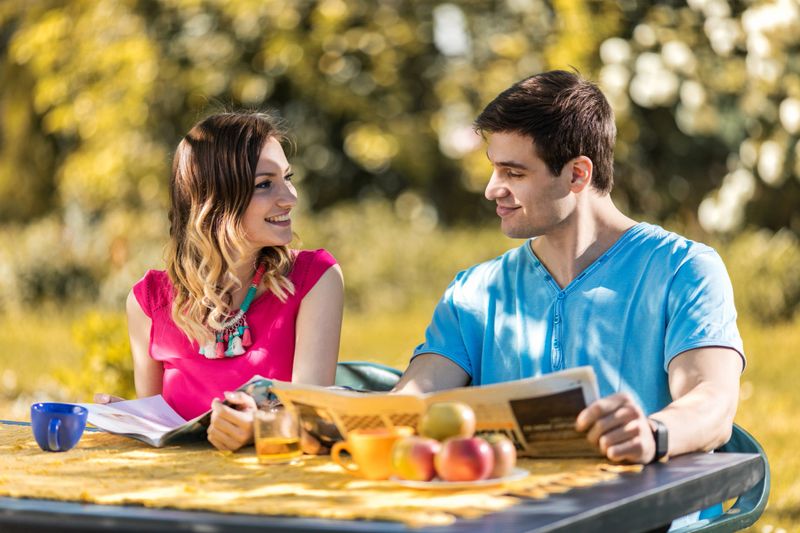 Smiling woman reading magazine and talking with her boyfriend who is reading newspaper while relaxing in their garden.