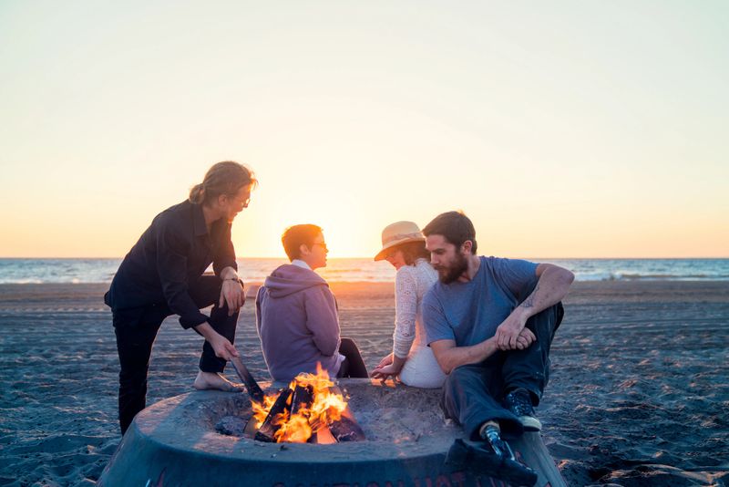 Group of young adult friends enjoying a fire on the beach as the sun sets. Los Angeles, America. October 2016