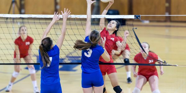High school girls volleyball match with players jumping at the net.