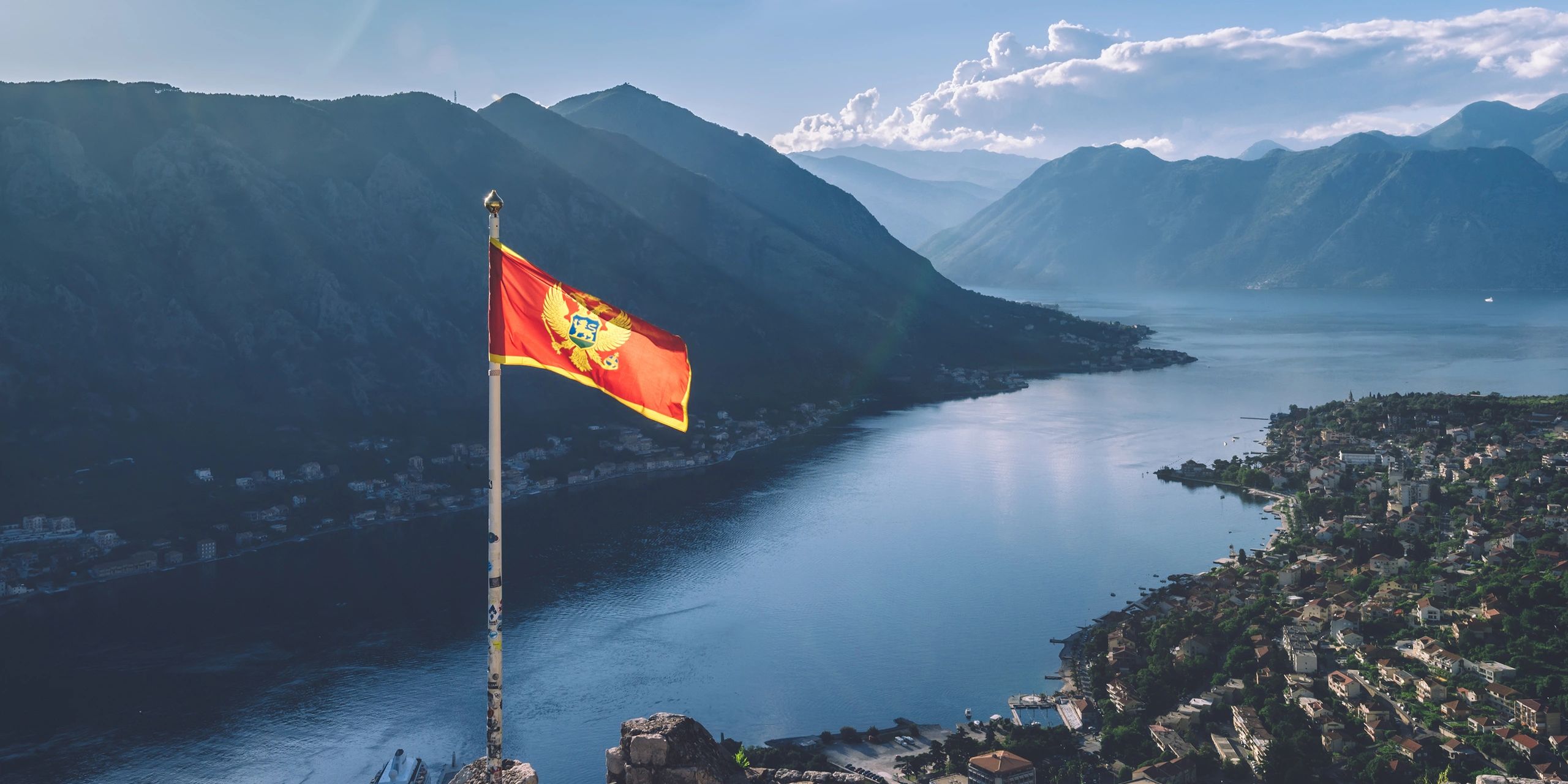 Montenegro flag waving over a scenic bay with mountains and town.