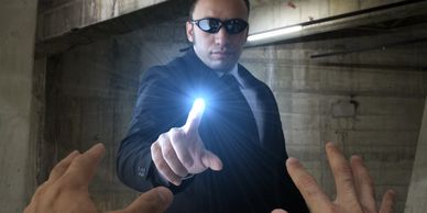 Man in black suit and sunglasses points glowing finger in dim concrete space.