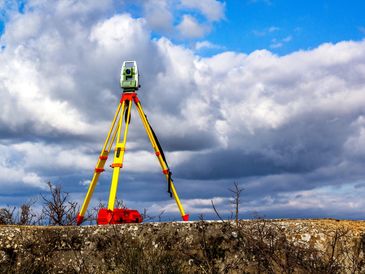 A surveying tripod stands against a cloudy sky on a stone surface.
