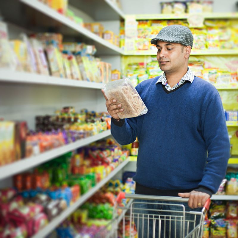 Image shot at supermarket in India in the month of November of an Indian young man doing shopping. He is holding and reading the label of a product near the shelves full of consumer products. One person, three quarter length, selective focus and square composition with copy space.
