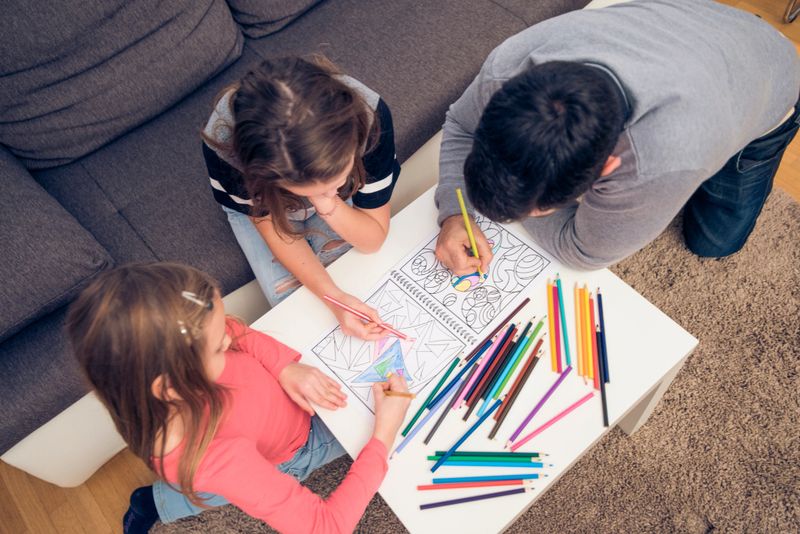 Father and two daughters are sitting in a living room. They are colouring books together.