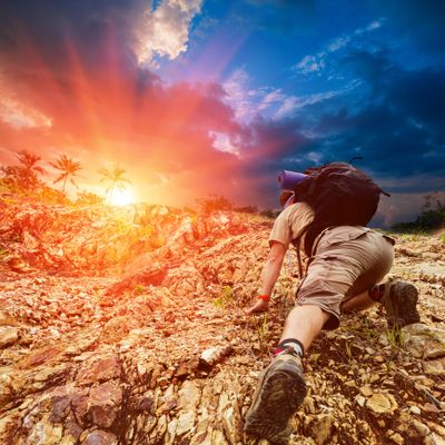 A hiker climbs rocky terrain towards a vibrant sunrise with palm trees in the background.