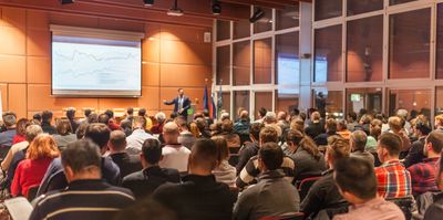 Audience attentively listens to a speaker presenting data on a large screen in a conference room.