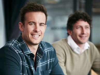 Two men smiling and sitting indoors in casual and business casual attire.