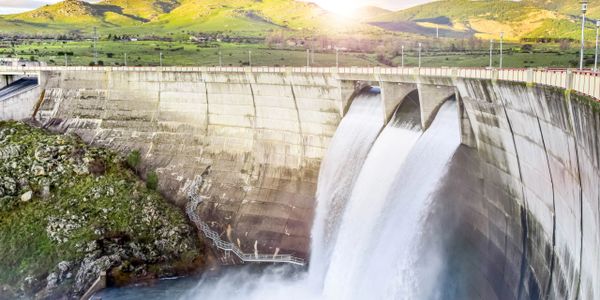 A dam releasing water with green hills and sun in the background.