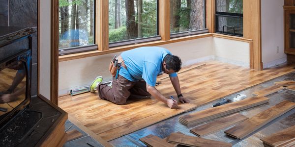 A man installs wooden flooring near large windows in a bright room.