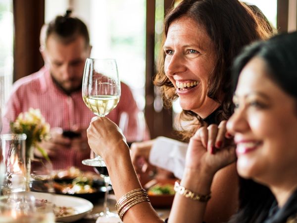 Two women laughing and enjoying wine at a dinner table with a man in the background.