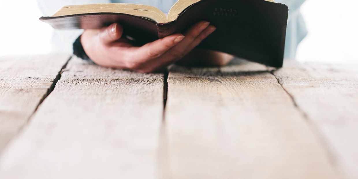 Person holding and reading an open Bible on a wooden table.