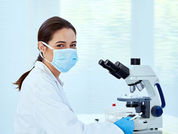 Female scientist in lab coat and mask working with a microscope.