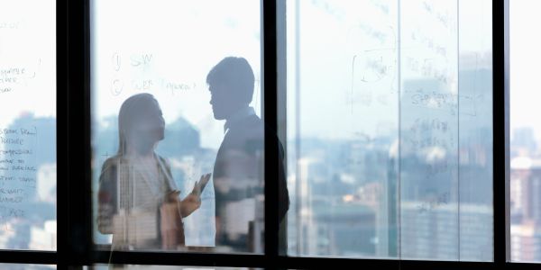 Silhouettes of two people discussing in an office with glass walls and city view.