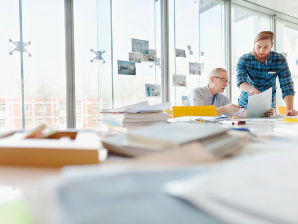 Two coworkers discuss documents in a bright office with large windows.