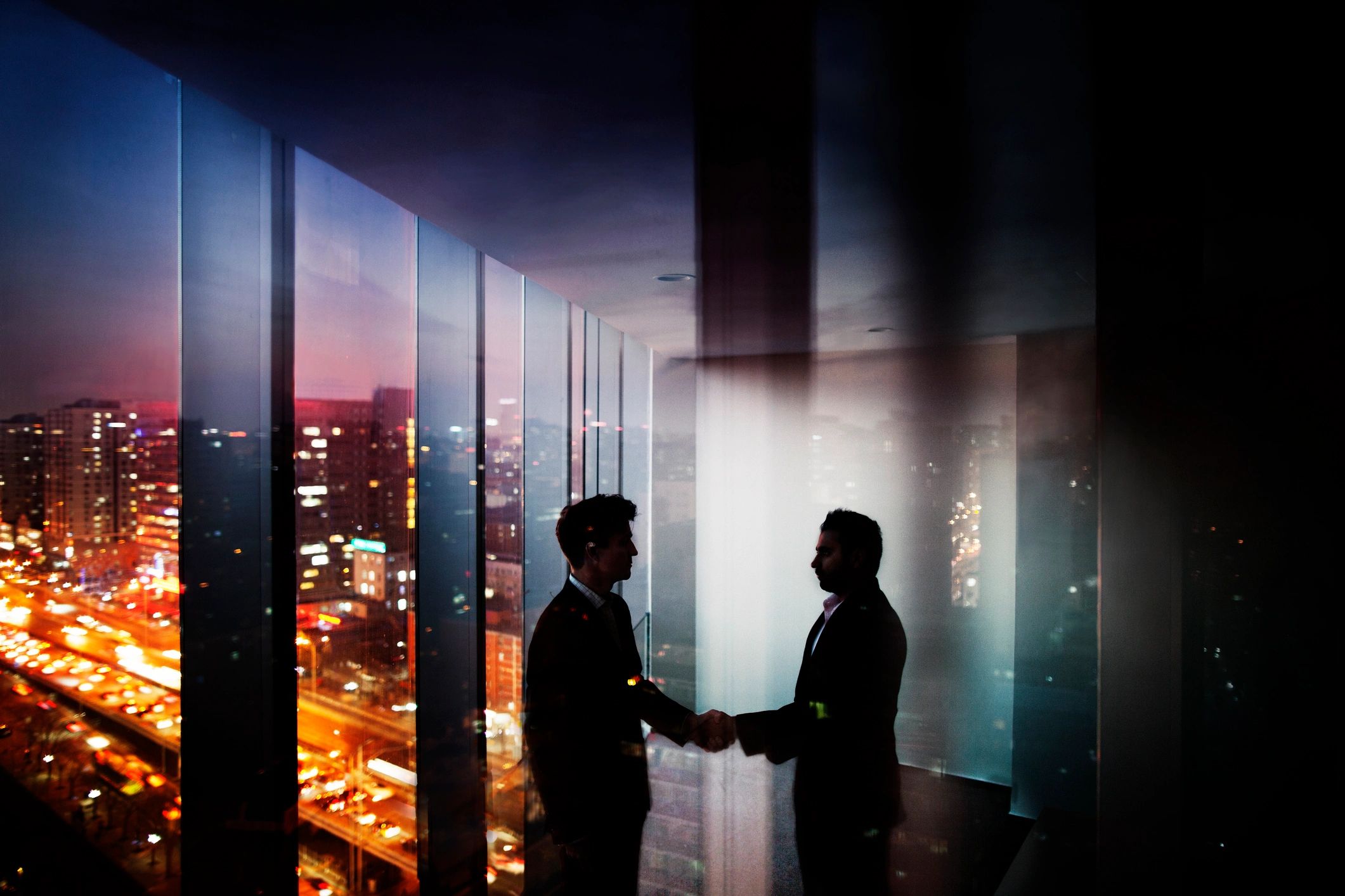 Two men shaking hands in an office with city lights outside at dusk.