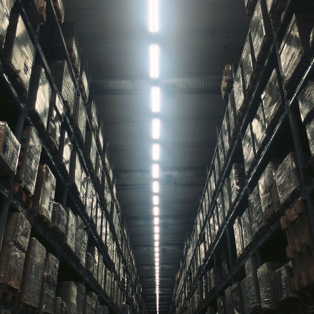 A long aisle in a warehouse filled with stacked pallets under bright overhead lights.