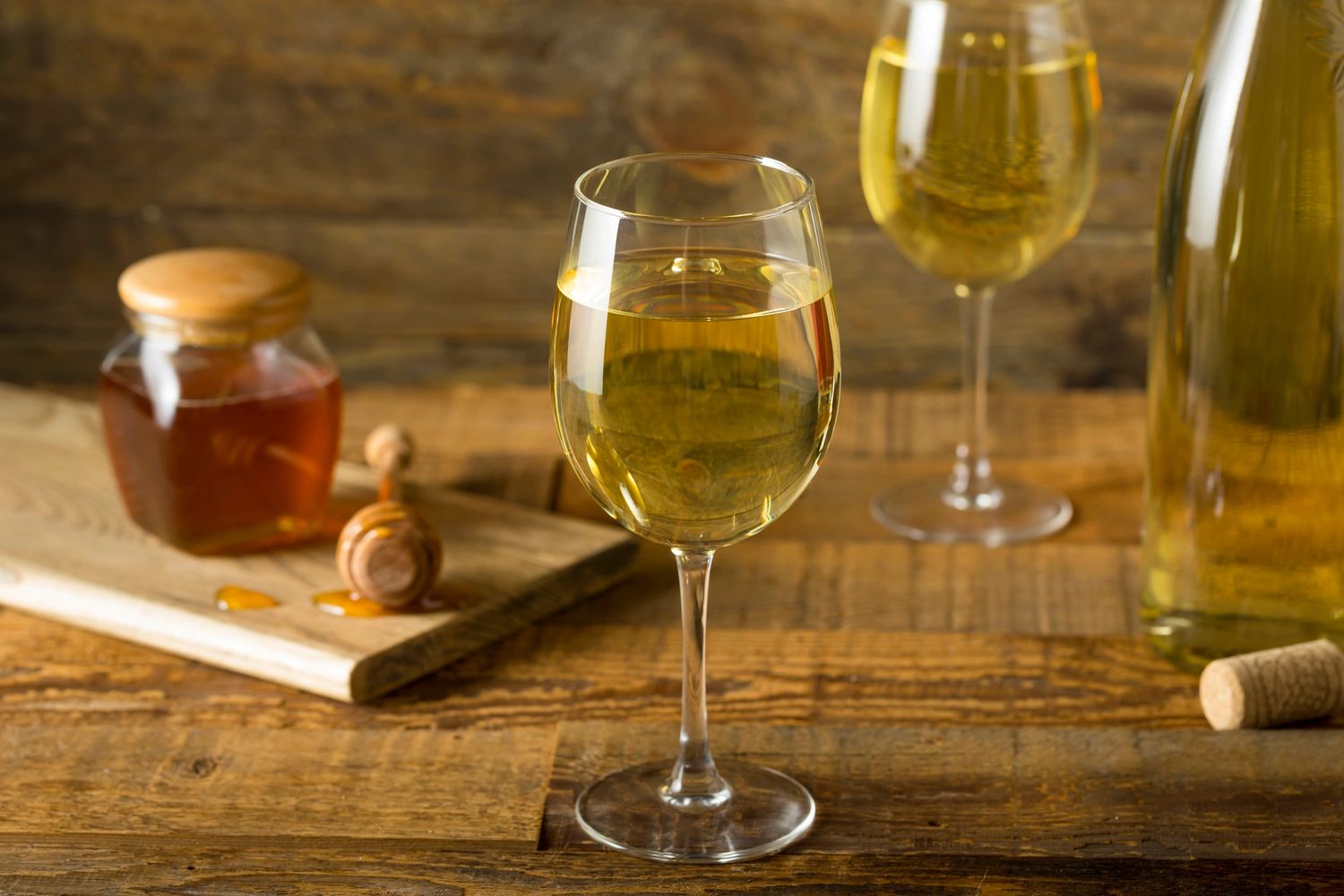 Glass of white wine with honey jar and cork on wooden table.