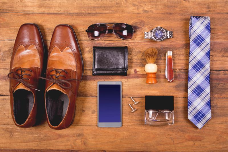 Variety of men's accessories organized in knolling arrangement.  Items include: dress shoes, tie, sunglasses, wallet, watch, shaving brush, cologne, and smart phone.  The items all lie on a wooden desk or table.   Men's personal accessories, clothing themes.  Father's Day.  Fashion.  Business.  Retail.
