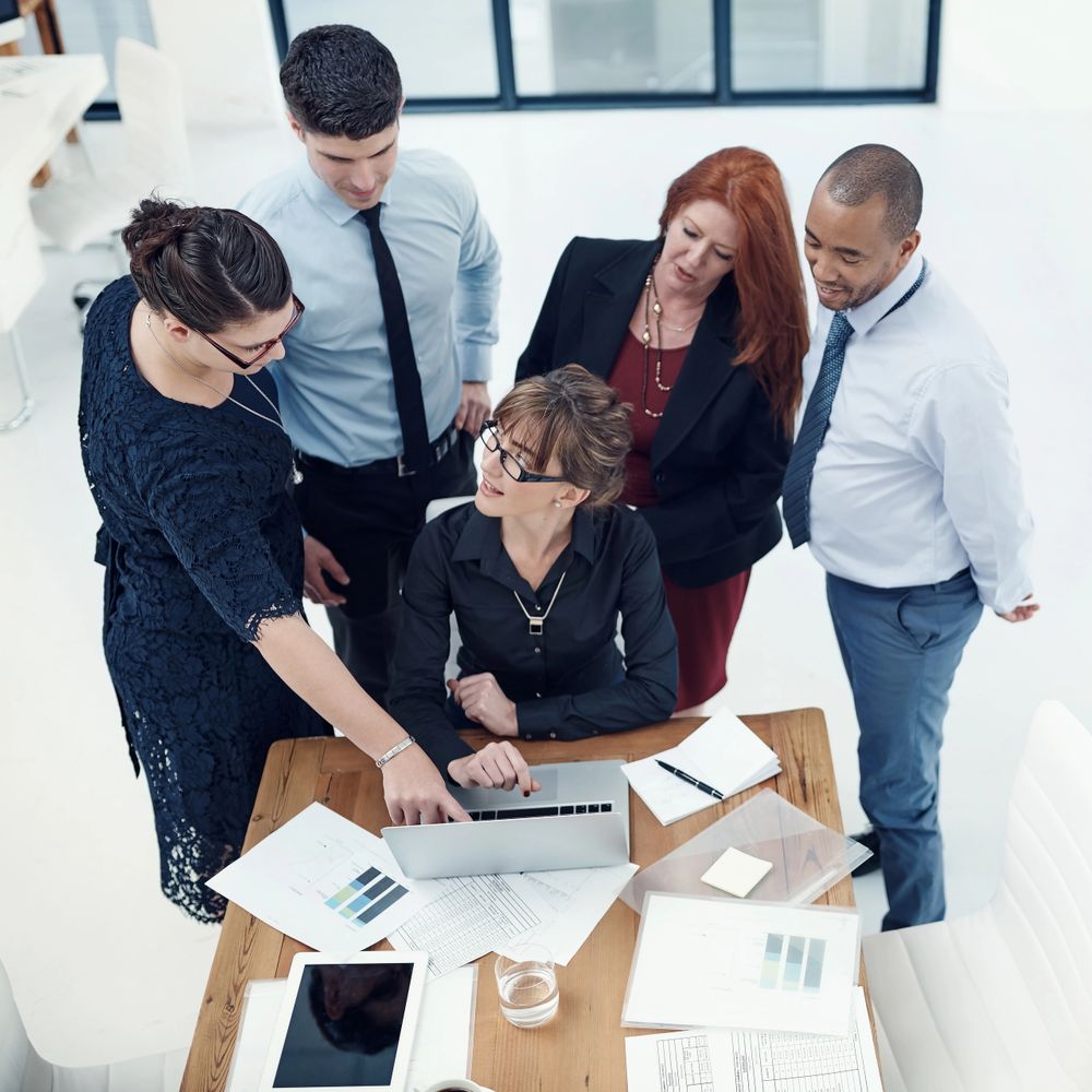 Five professionals collaborating around a laptop at a wooden table in a modern office.