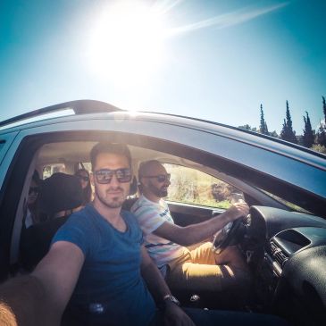 Two men in a car on a sunny day, one driving and one taking a selfie.
