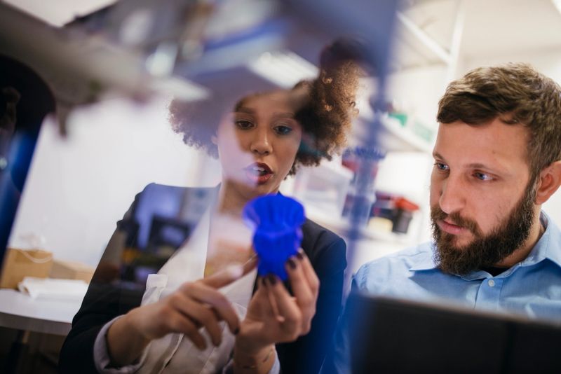 Close up shot of 3D printer printing 3D objects. Two workers are observing the process. Women is holding 3D object in her hand.