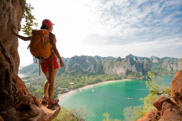 Hiker with backpack stands on a cliff overlooking a turquoise bay and green mountains.
