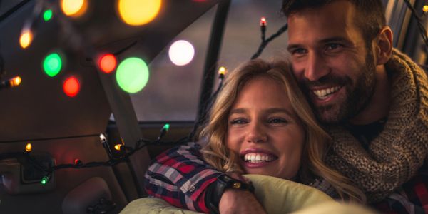 Couple cuddling in a car decorated with colorful string lights, wrapped in a blanket.