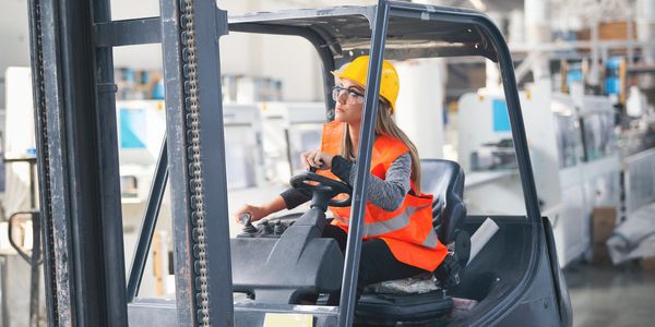 Woman operating a forklift in a warehouse, wearing safety gear.