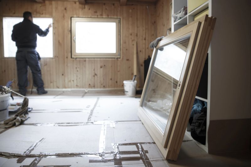 Worker in the background installing new, three pane wooden windows in an old wooden house, with a new window in the foreground. Home renovation, sustainable living, energy efficiency concept.