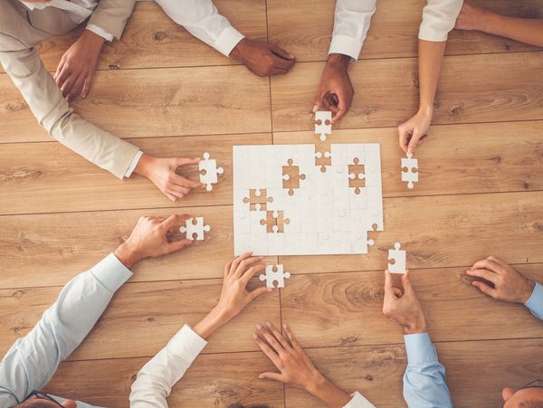 People collaborating on assembling a white puzzle on a wooden table.
