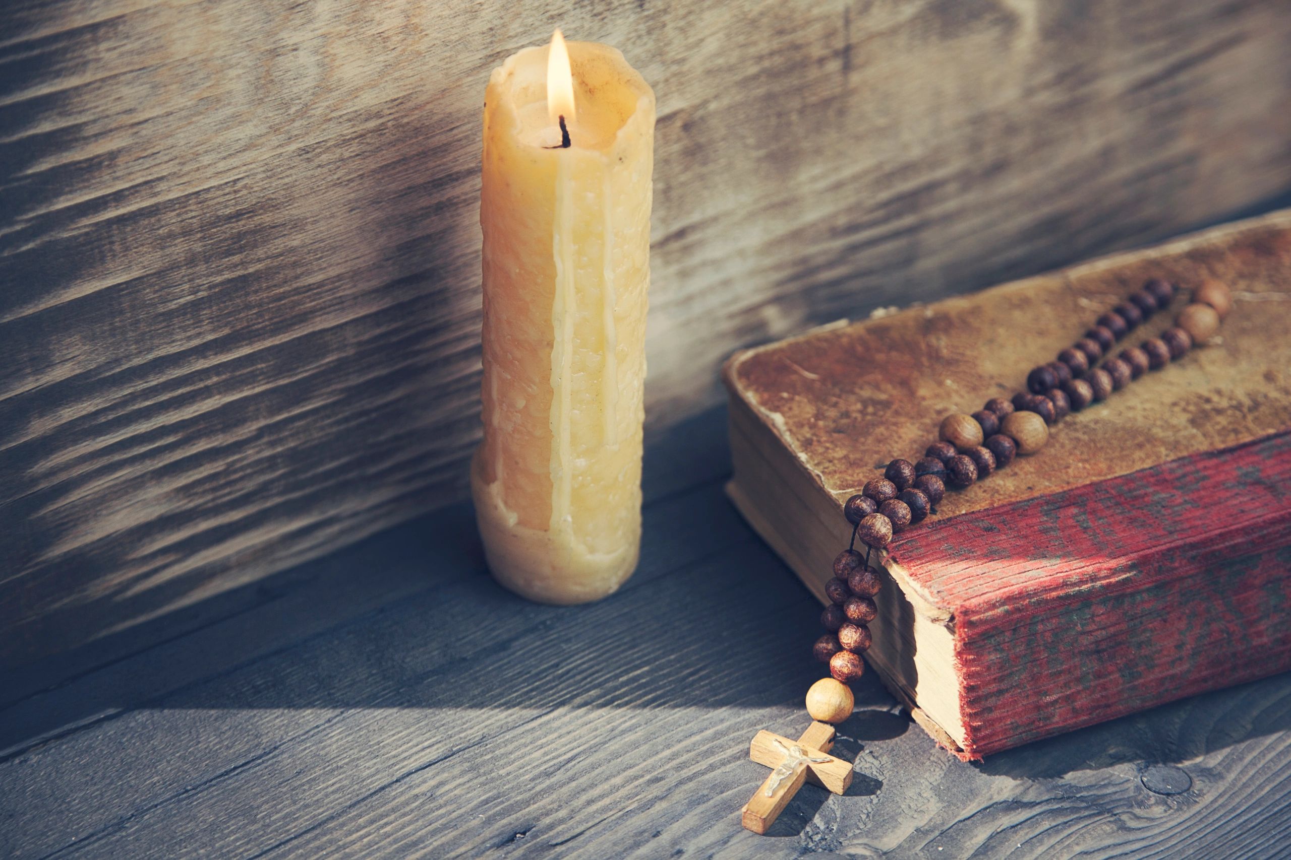A lit candle next to an old book with a wooden rosary.