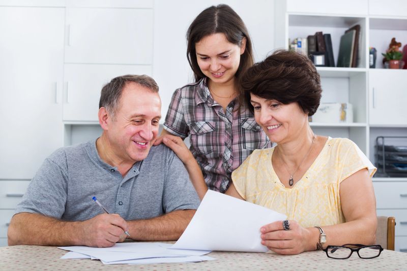 Smiling adult daughter helping with documents to her mature parents at a home. Focus on both mature persons