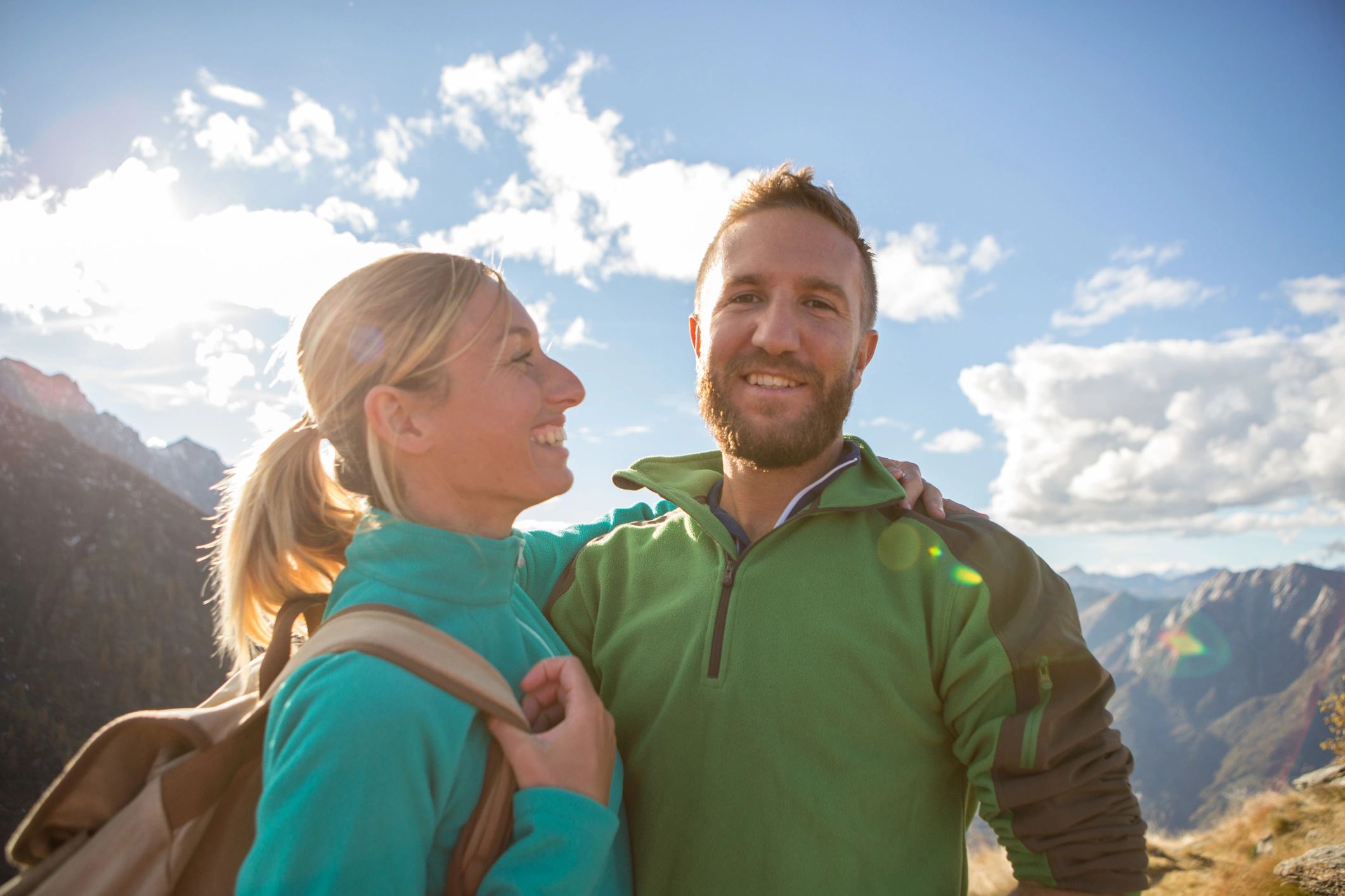 Couple smiling outdoors with mountains and sky in background.