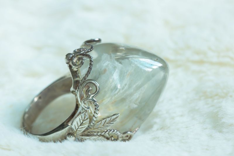 A quartz pebble is formed with tiny silica crystals inside. This well-polished pebble, is on a silver ring that decorated with engraved tree leaves. The silver ring is placed on soft white wool.