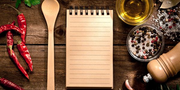 Empty notepad surrounded by spices, wooden spoon, and oil on a wooden table.